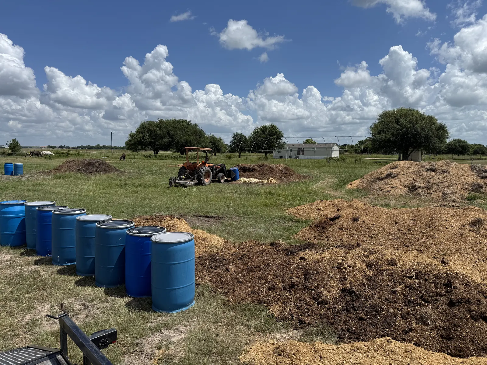 Blue collection drums beside long compost windrows of food waste and wood chips on a Texas farm, with horses grazing in the background